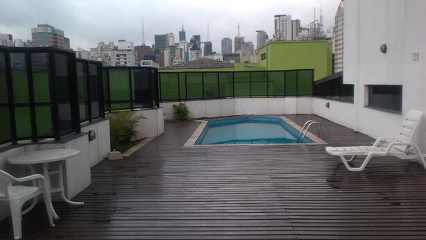 Un área de piscina en la azotea con vistas a un horizonte urbano, terraza de madera y sillones de descanso blancos.