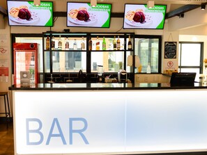A bar area with a counter, multiple screens displaying desserts, a variety of liquor bottles, and a menu board. - ibis Styles Rio Preto Monte Líbano (São José do Rio Preto)