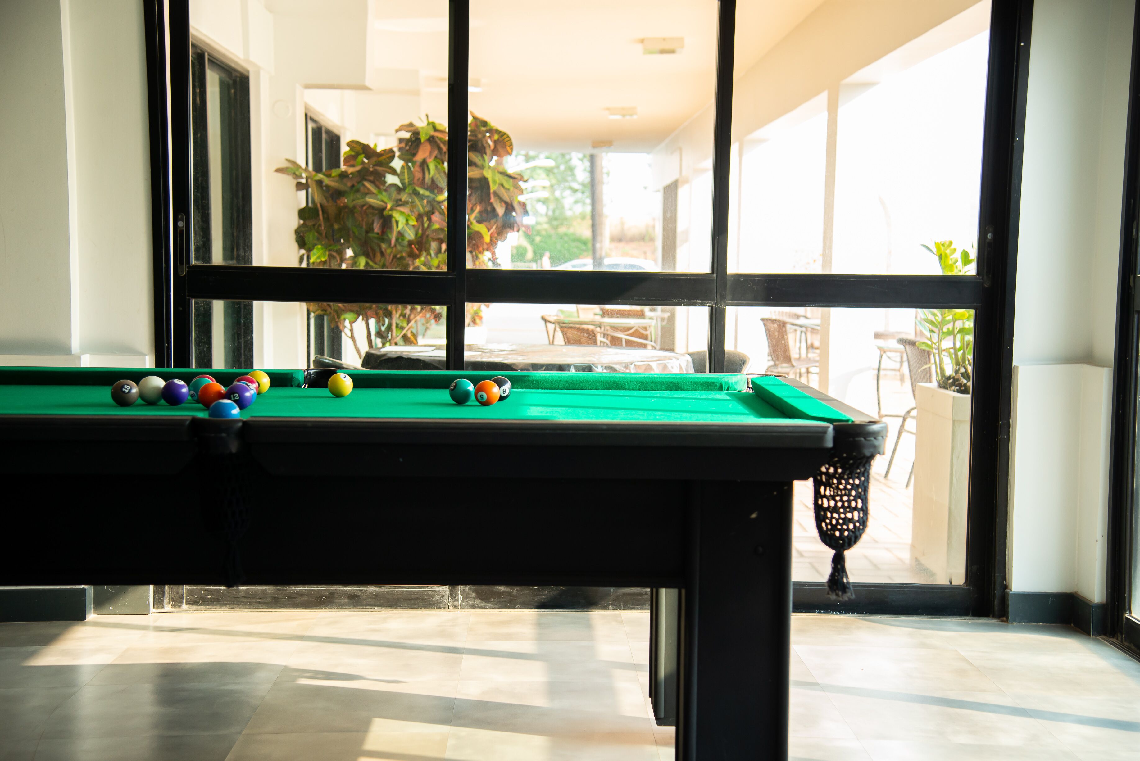 Una mesa de billar con bolas, una ventana y vistas a un espacio exterior con sillas y plantas.