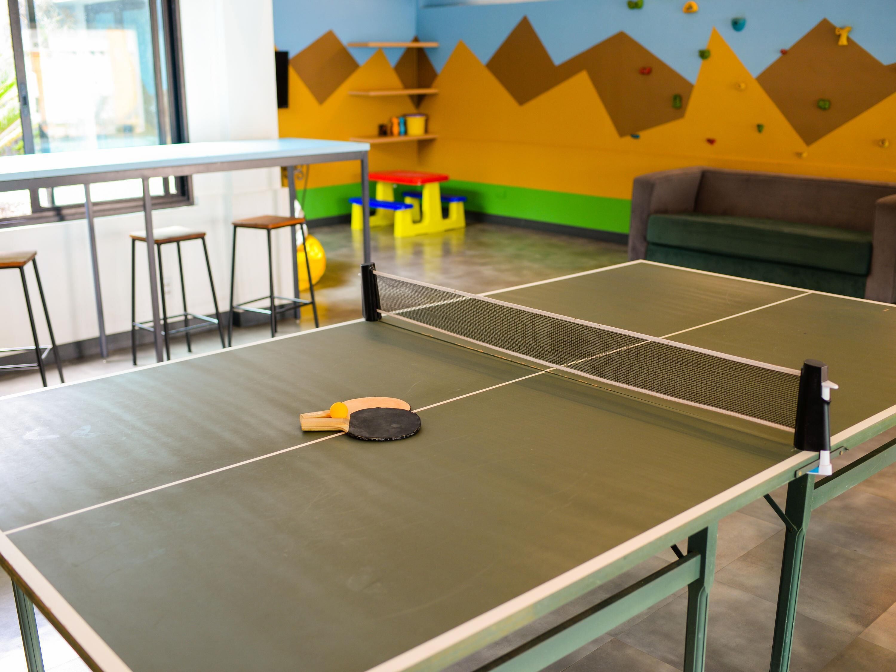 Tafeltennistafel met net, stoelen en een bijzettafeltje in een kamer met kleurrijke wanden en een bank.