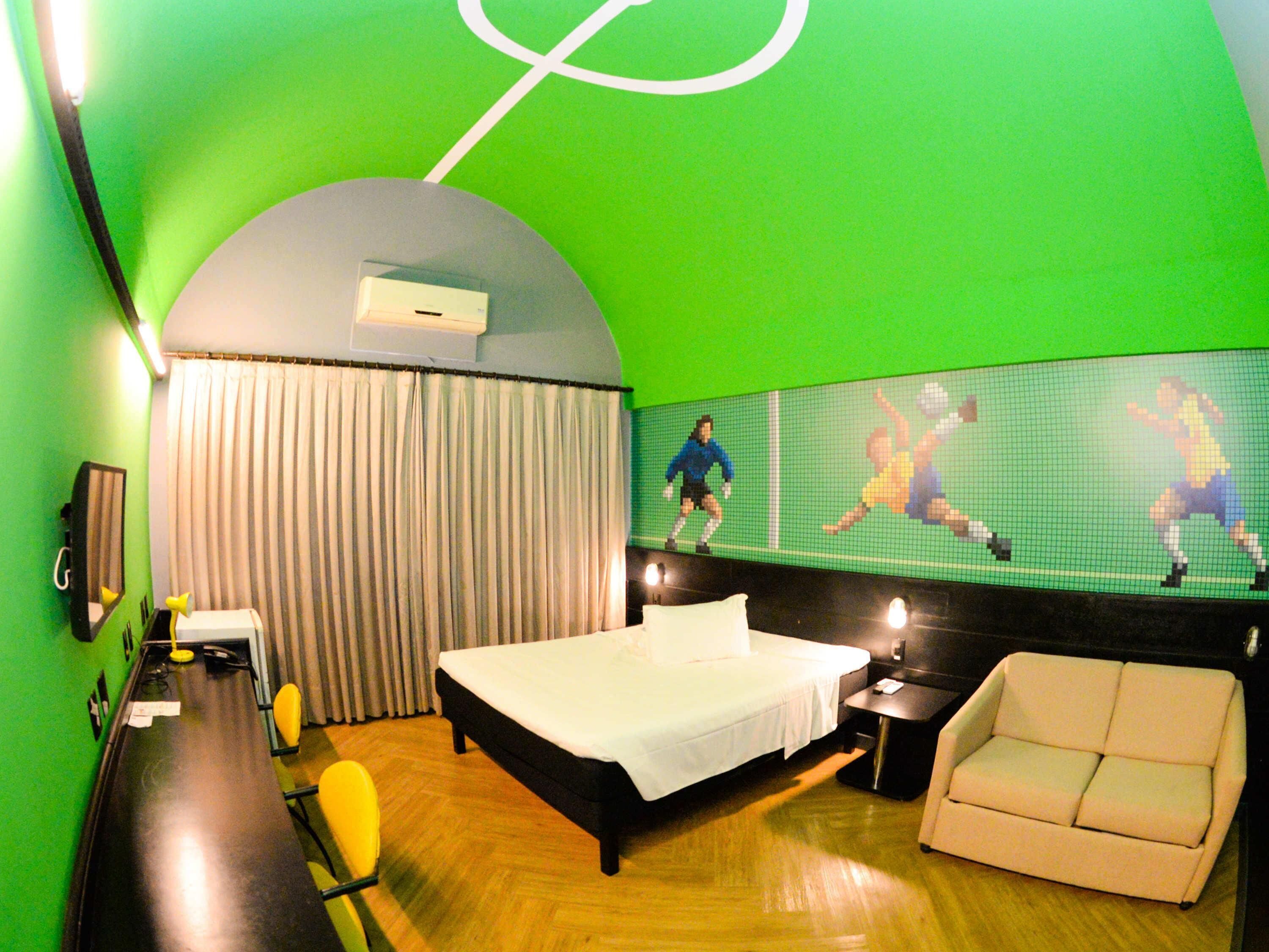 A hotel room with a bed, a sofa, a desk, and a large digital screen displaying a sports game.