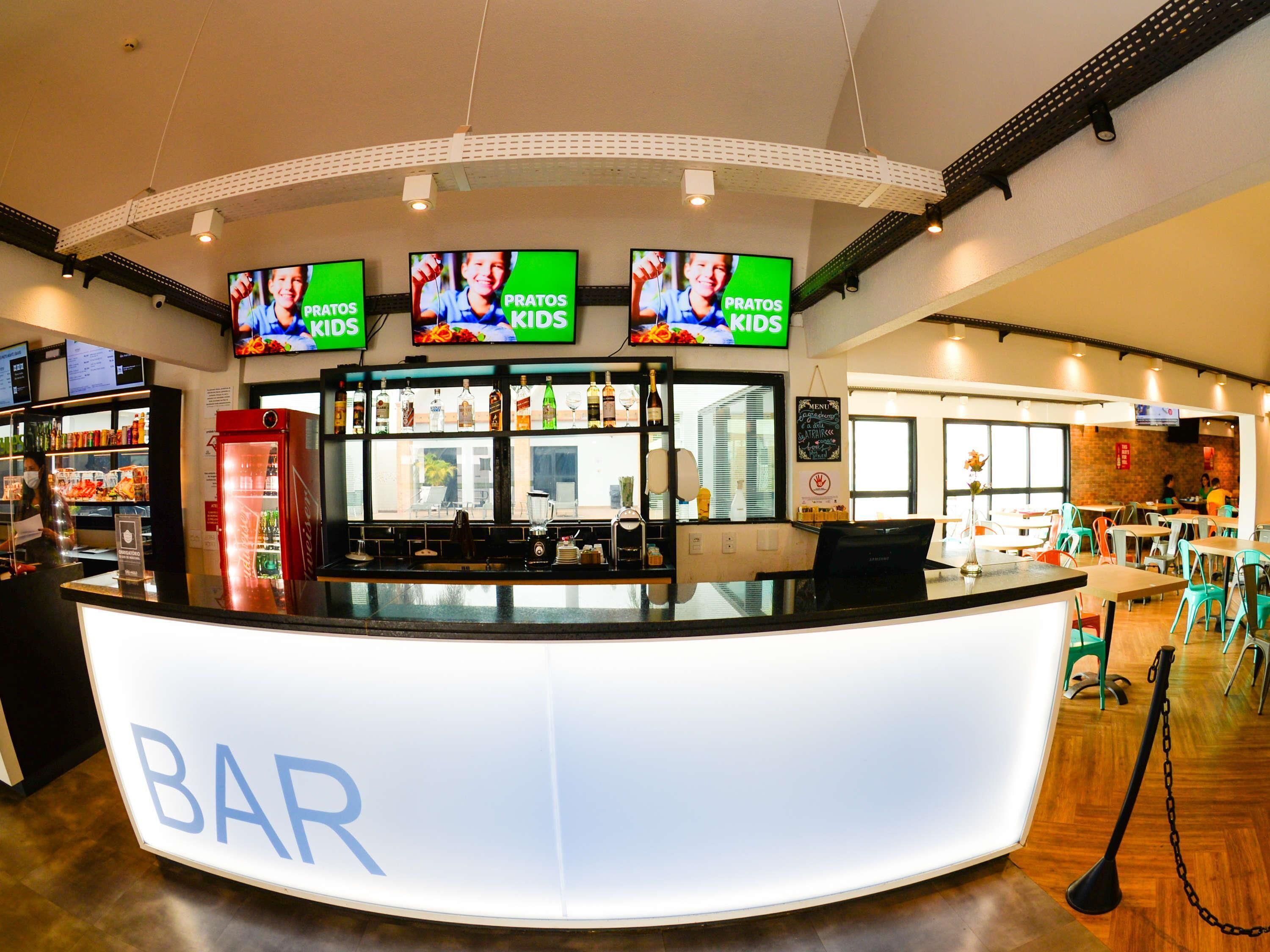 A bar area with a counter, multiple screens displaying content, a refrigerator, and a seating area with tables and chairs.