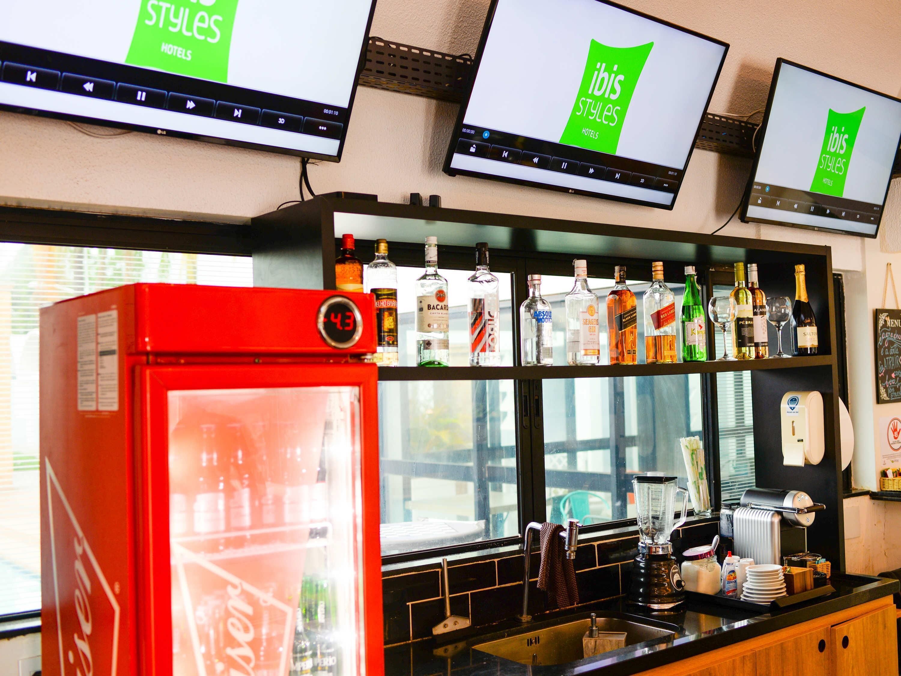 Et barområde med flere skærme, der viser 'ibis Styles Hotels' branding, et udvalg af spiritusflasker, et rødt køleskab og en kaffemaskine.