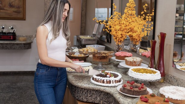 A woman in a white top and blue jeans serving food at a buffet with various cakes and pastries.