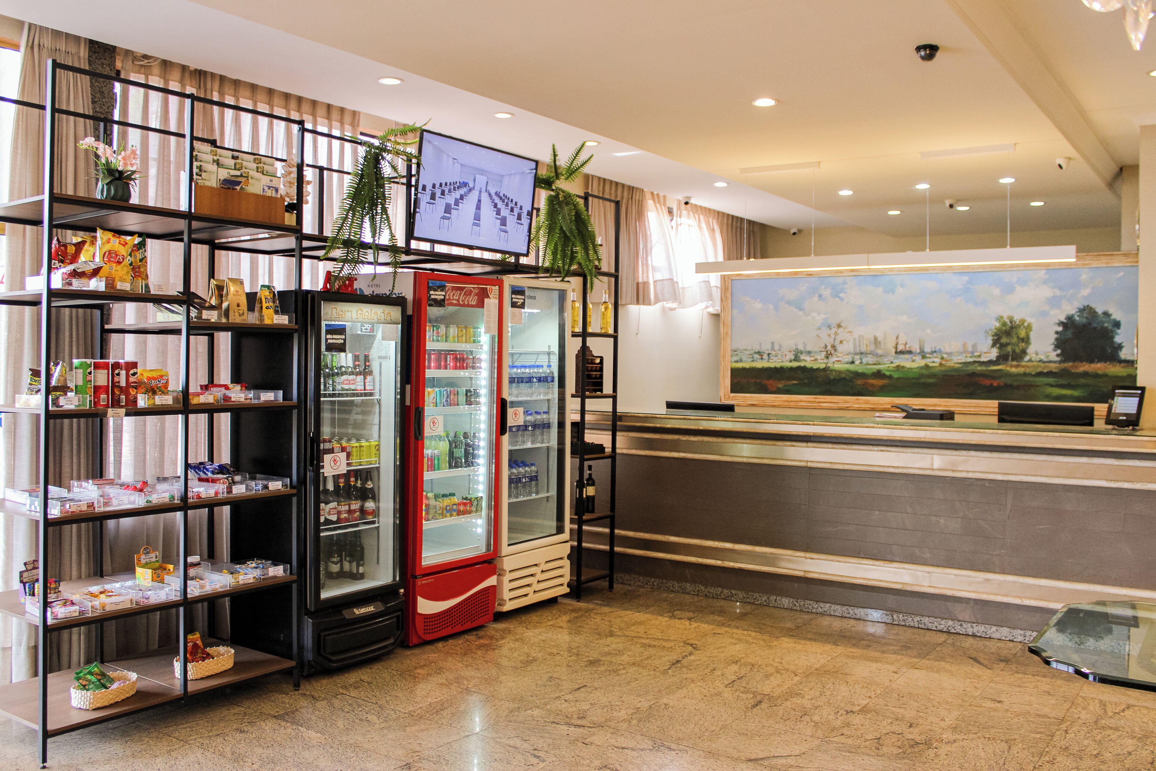 A hotel lobby with a reception desk, a Coca-Cola refrigerator, a television mounted on the wall, and a large painting of a landscape.