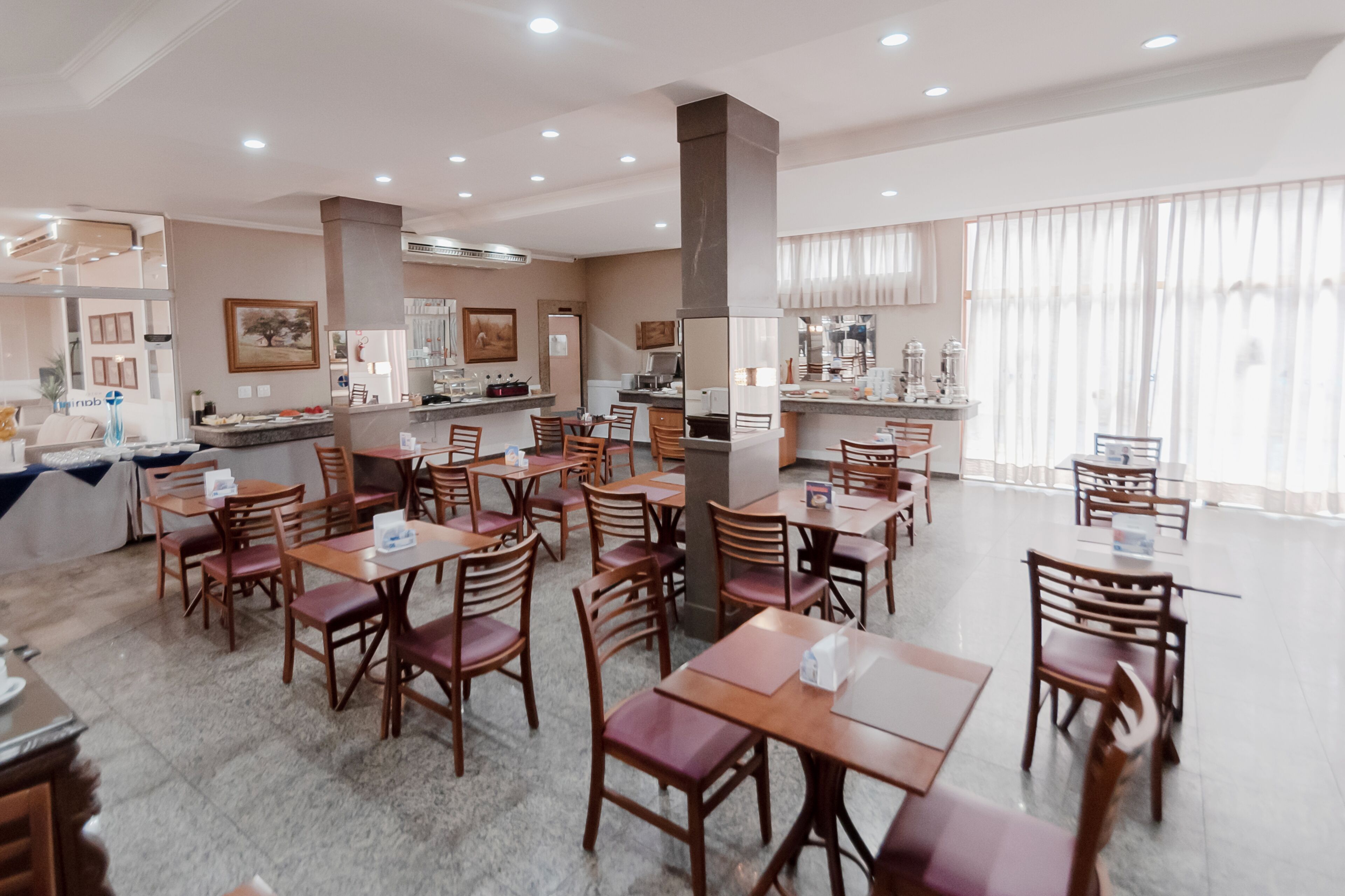 Un grand espace dédié à la salle à manger, avec de nombreuses tables et chaises en bois, un pilier central et, au fond, un coin cuisine.