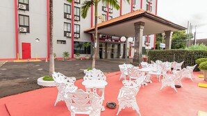 A hotel exterior with a red and white facade, outdoor seating area with white chairs and tables, and a palm tree. - Hotel Dan Inn Araraquara By Nacional Inn  (Araraquara)