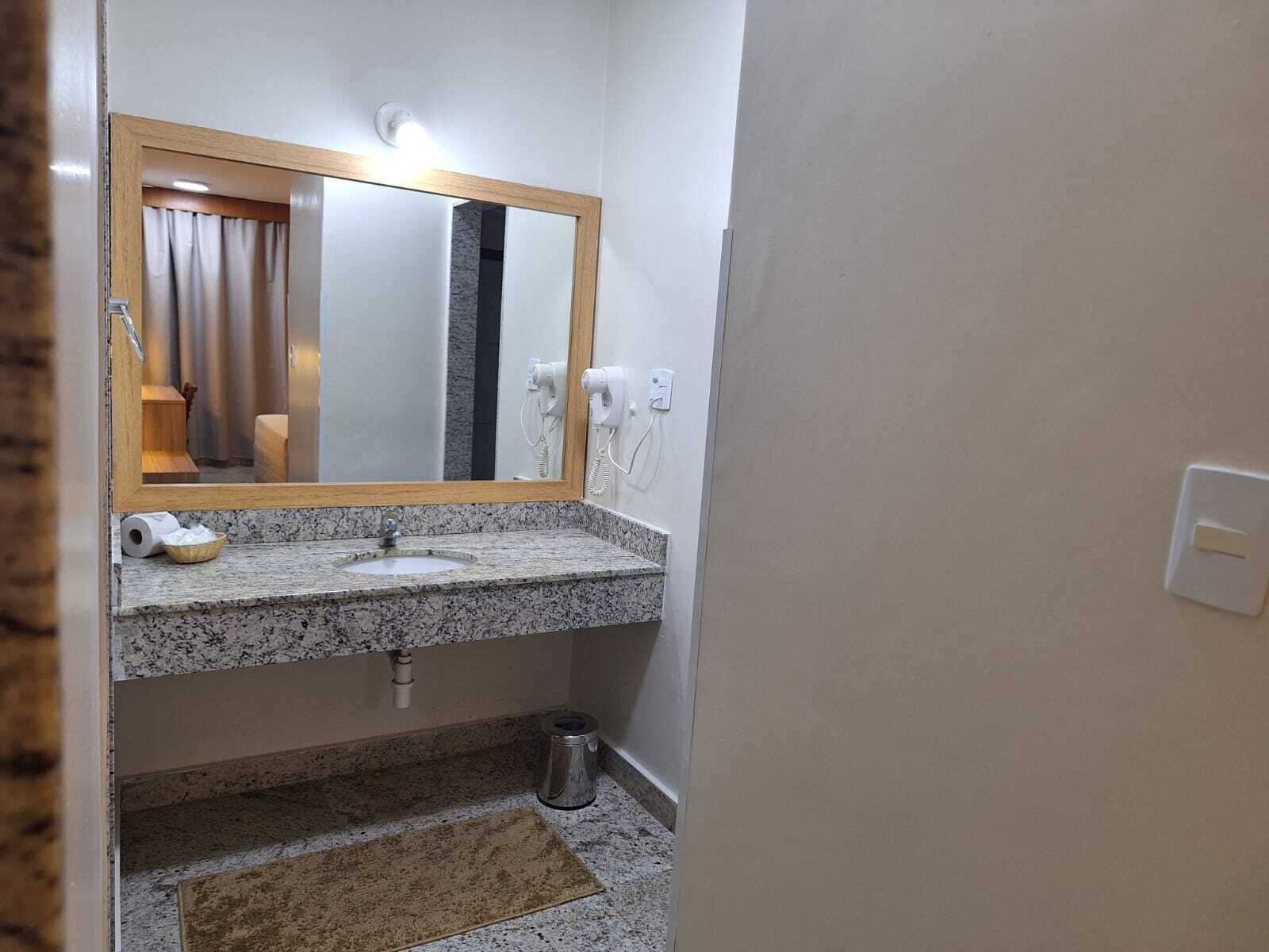 A bathroom with a granite countertop sink, a mirror with a wooden frame, a hairdryer, and a trash can.