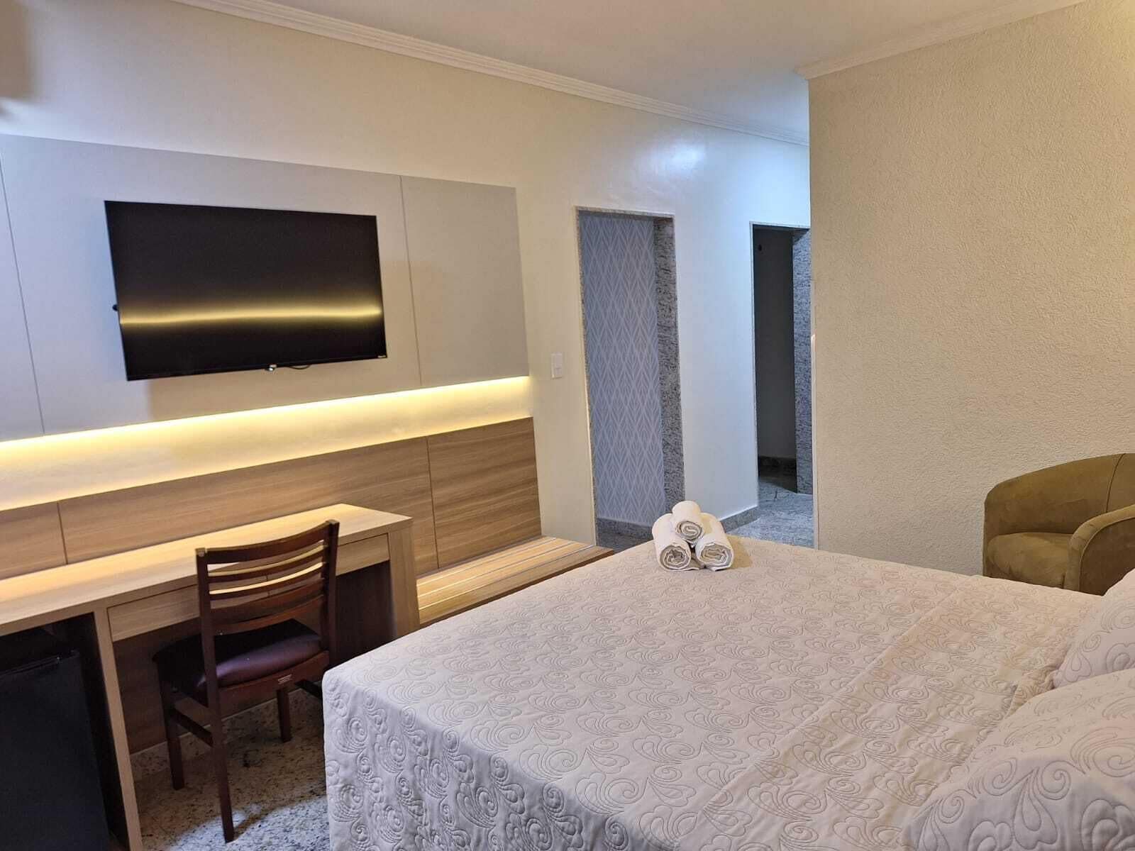 A hotel room with a bed, a desk with a chair, a television mounted on the wall, and a bathroom visible through an open door.