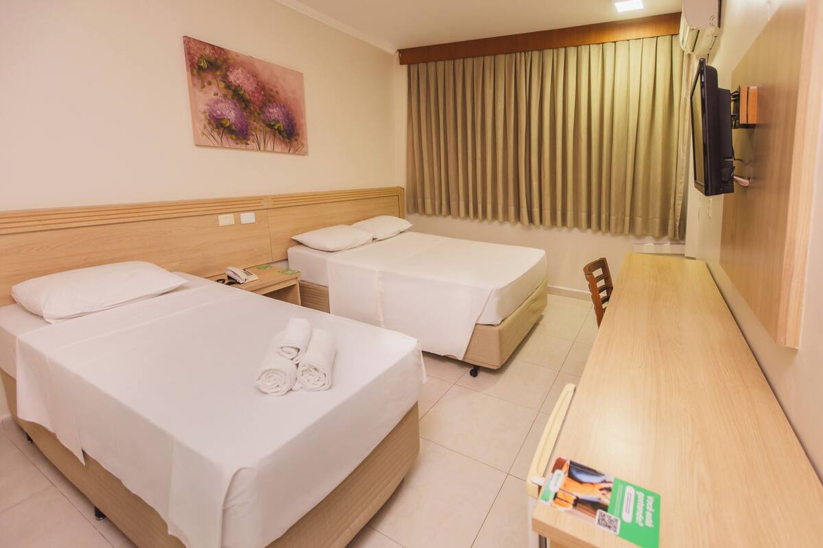 A hotel room with two beds, a wooden desk, a painting on the wall, and a curtain-covered window.