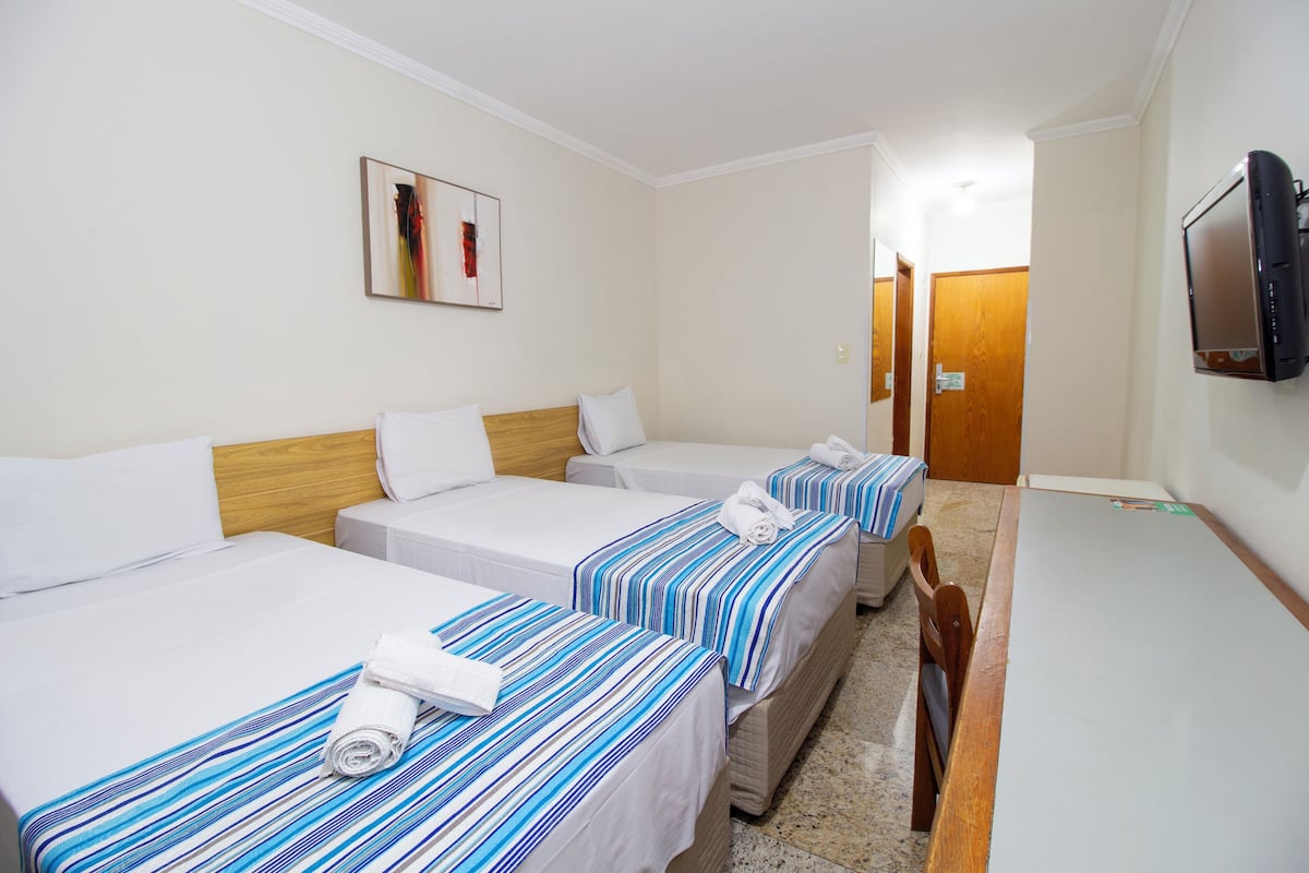 A hotel room with three single beds, each with a striped bedspread and rolled towels. A television is mounted on the wall, and a wooden door is visible in the background.