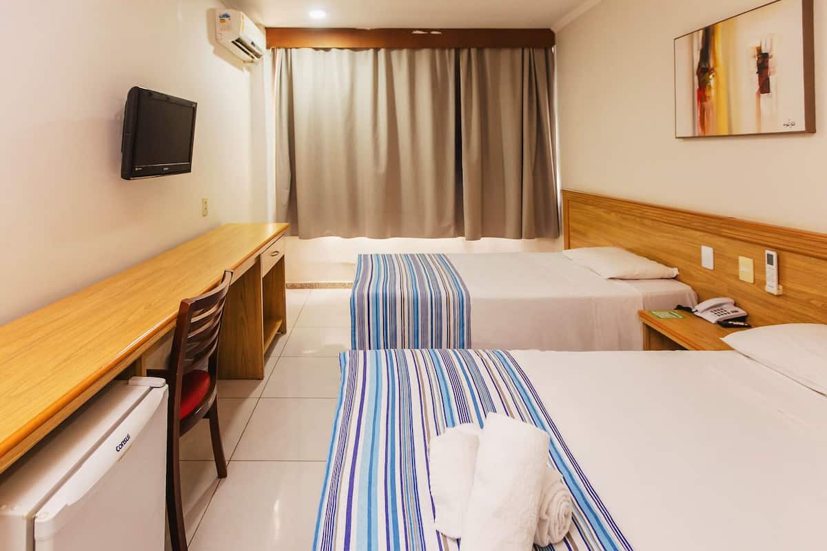 A hotel room with two beds, a desk, a chair, a refrigerator, a TV, and a window with curtains.