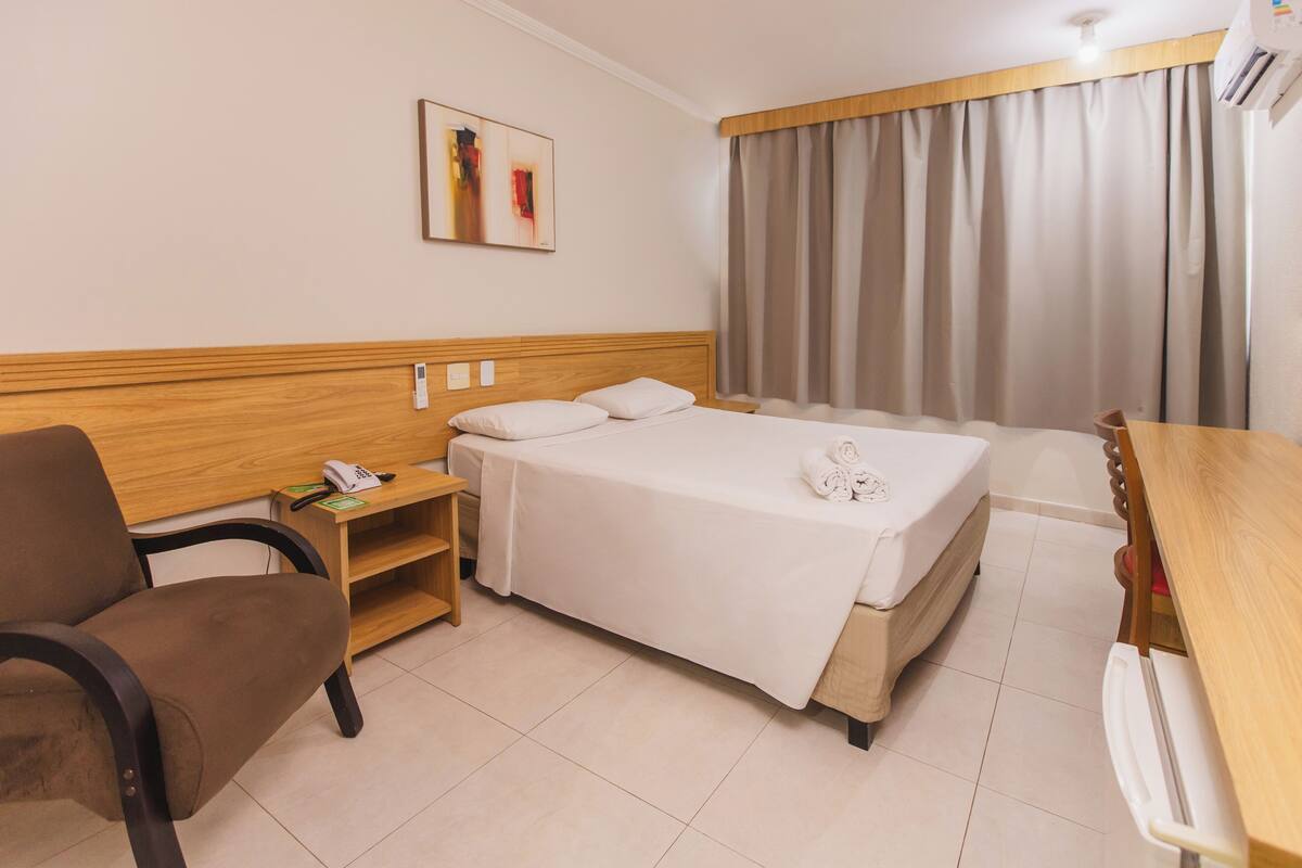 A hotel room with a bed, a desk, a chair, a bedside table, and a wall-mounted telephone.