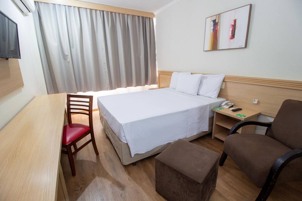 A hotel room with a bed, a desk, a chair, a bedside table with a phone, and a painting on the wall.