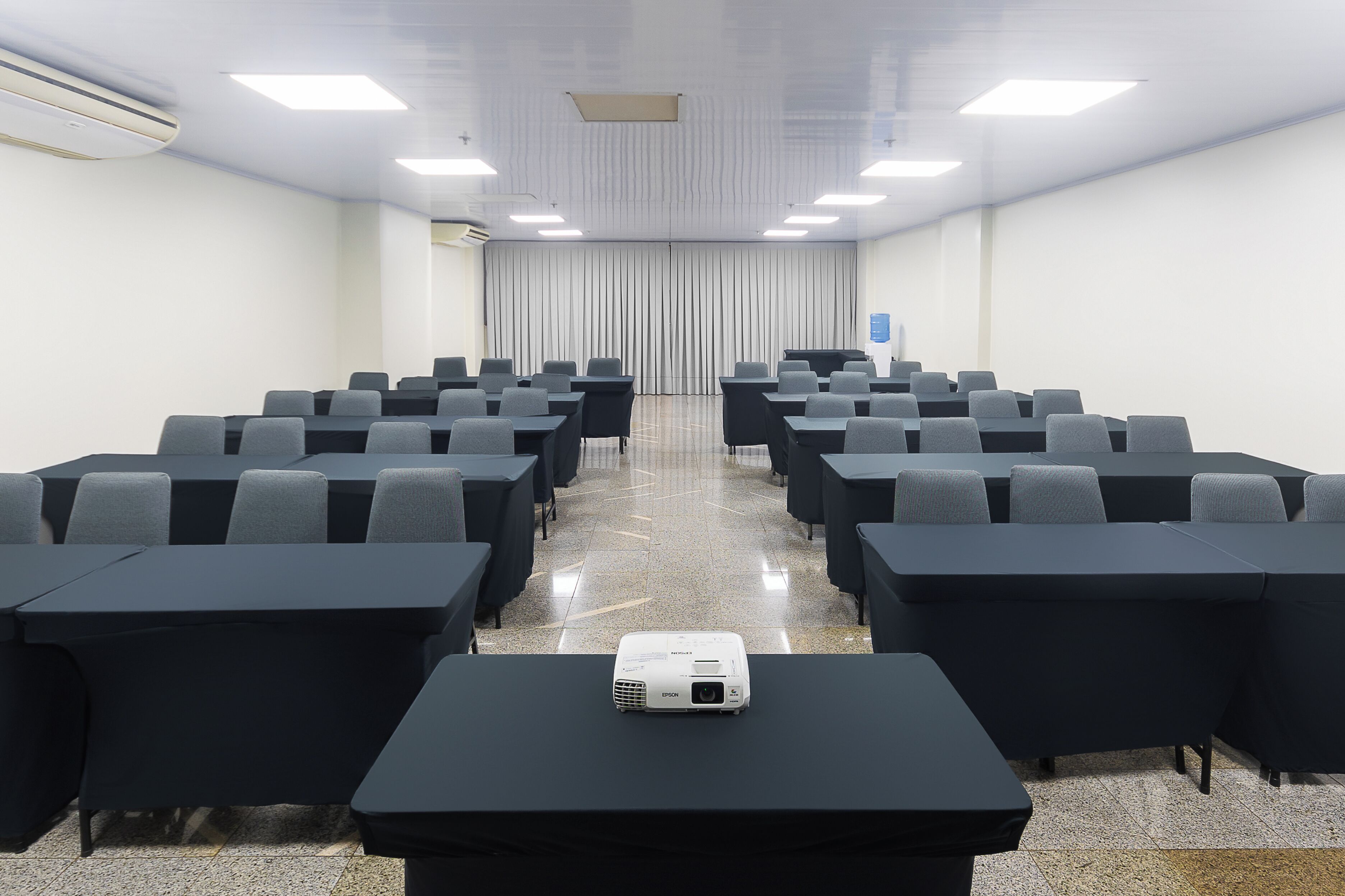 Un salón de conferencias con filas de mesas y sillas, un proyector sobre la mesa delantera y una cortina al fondo.