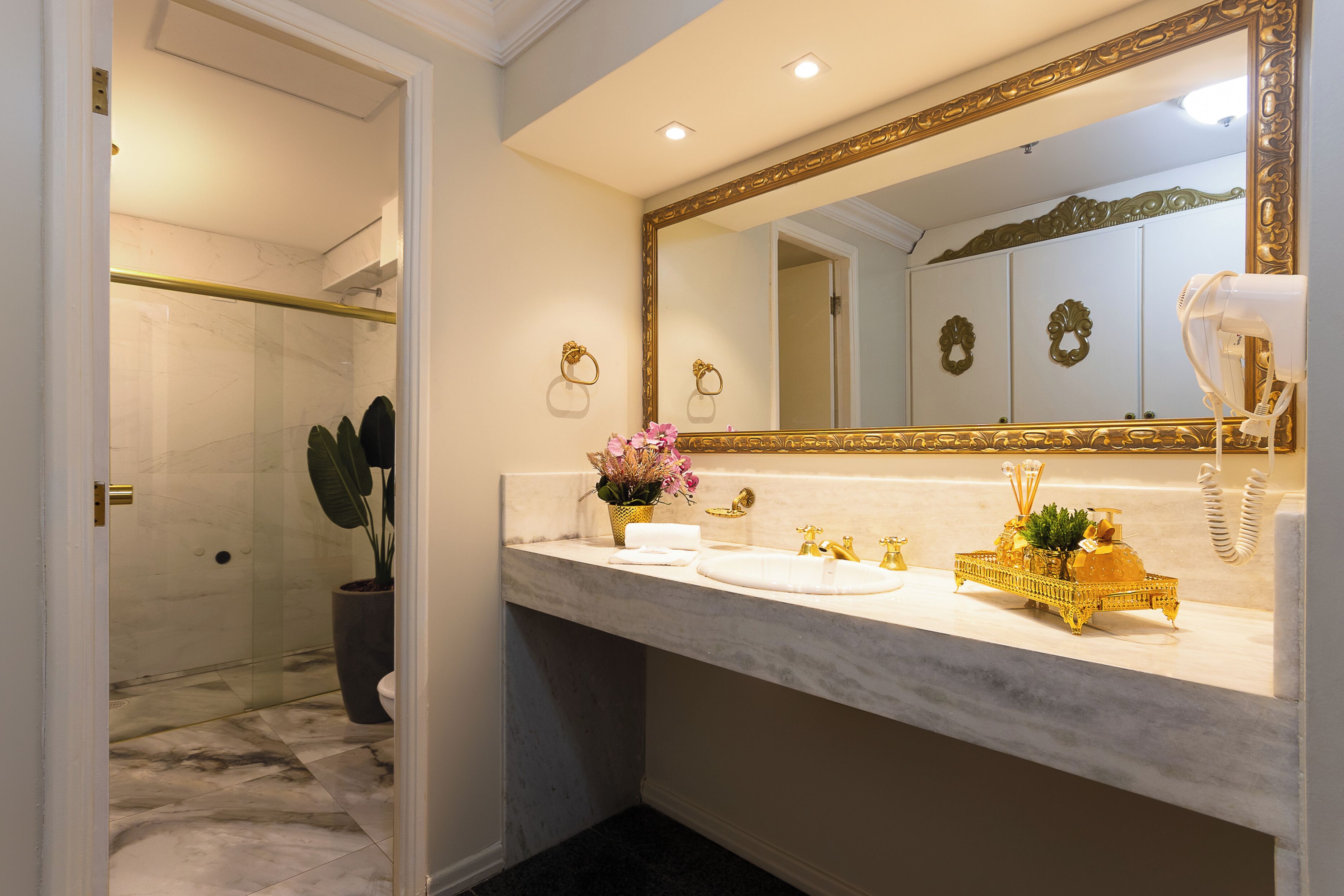 Une salle de bain avec un lavabo en marbre, des robinets dorés, un grand miroir et une plante en pot.