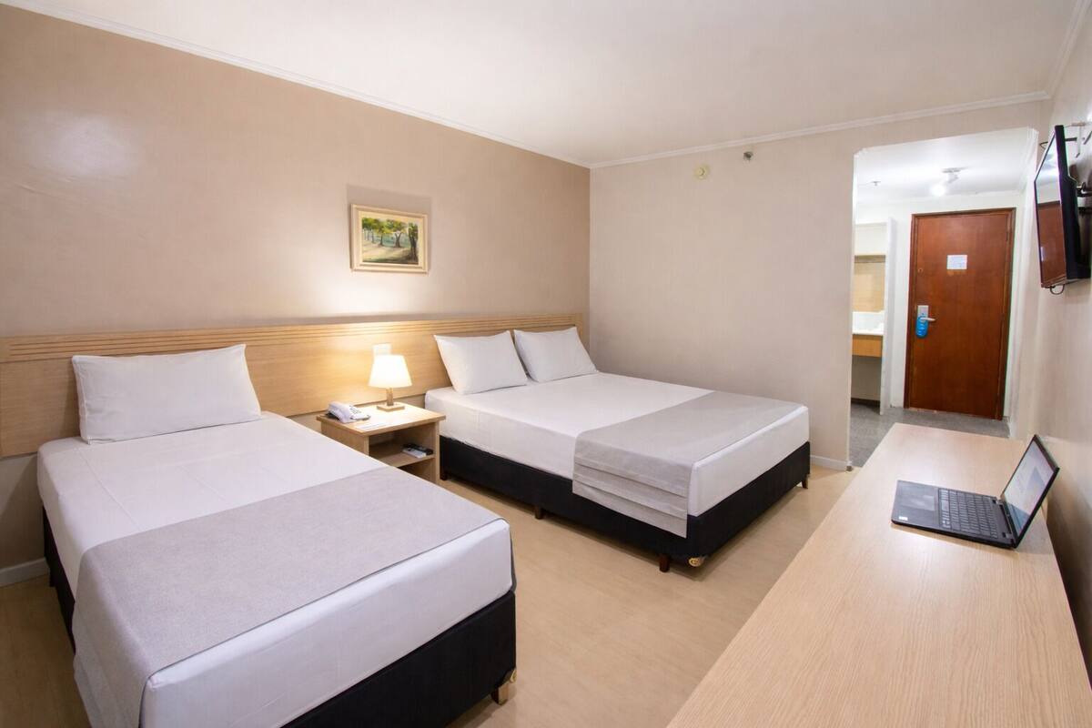Ein Hotelzimmer mit zwei Betten, einem Schreibtisch mit einem Laptop, einer Nachttischlampe und einem Bild an der Wand.