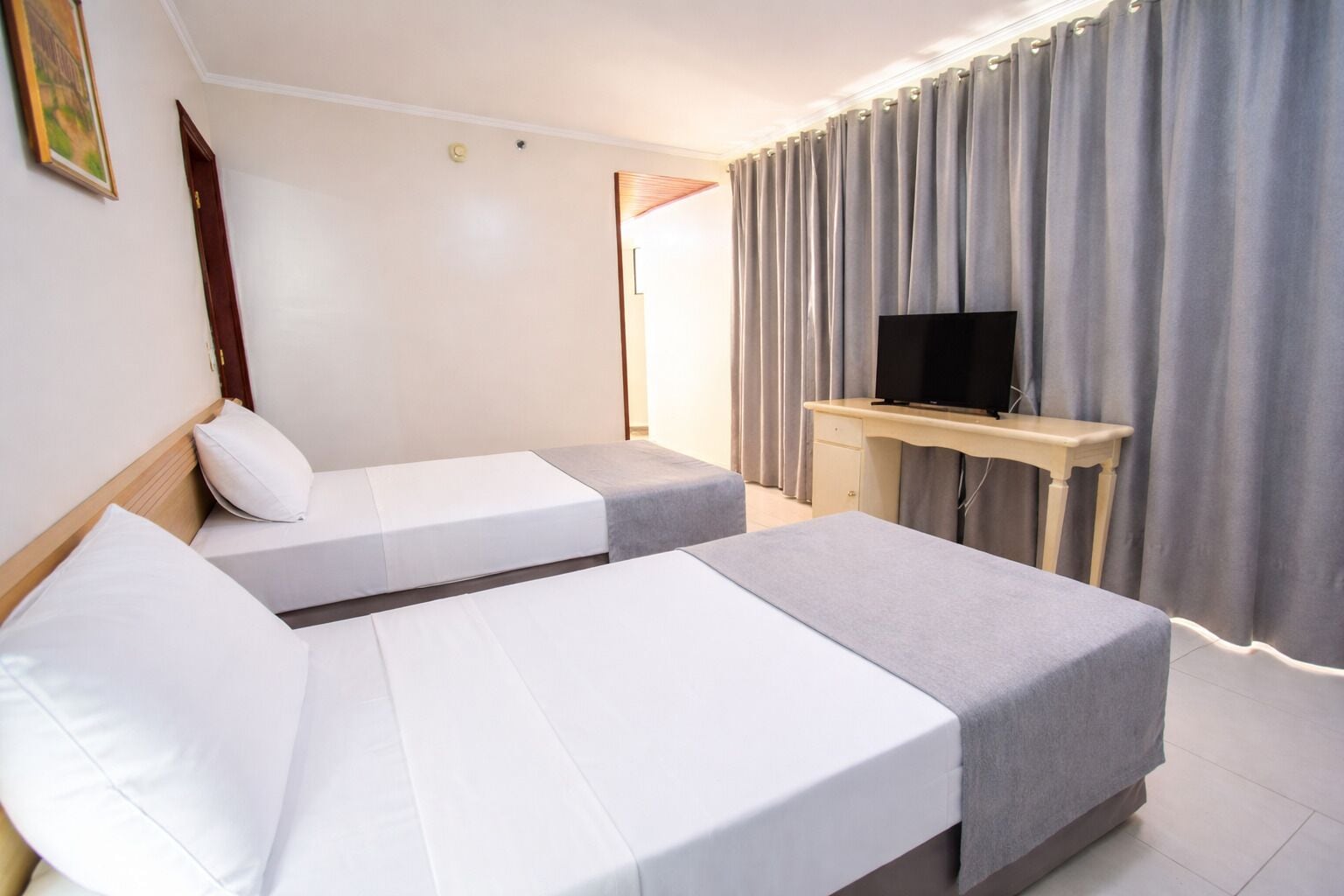 Una habitación de hotel con dos camas, un escritorio con televisor y una ventana con cortinas.