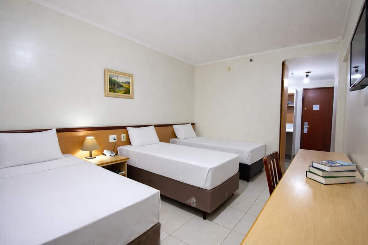Ein Hotelzimmer mit drei Betten, einem hölzernen Schreibtisch, einer Lampe und einem Gemälde an der Wand.