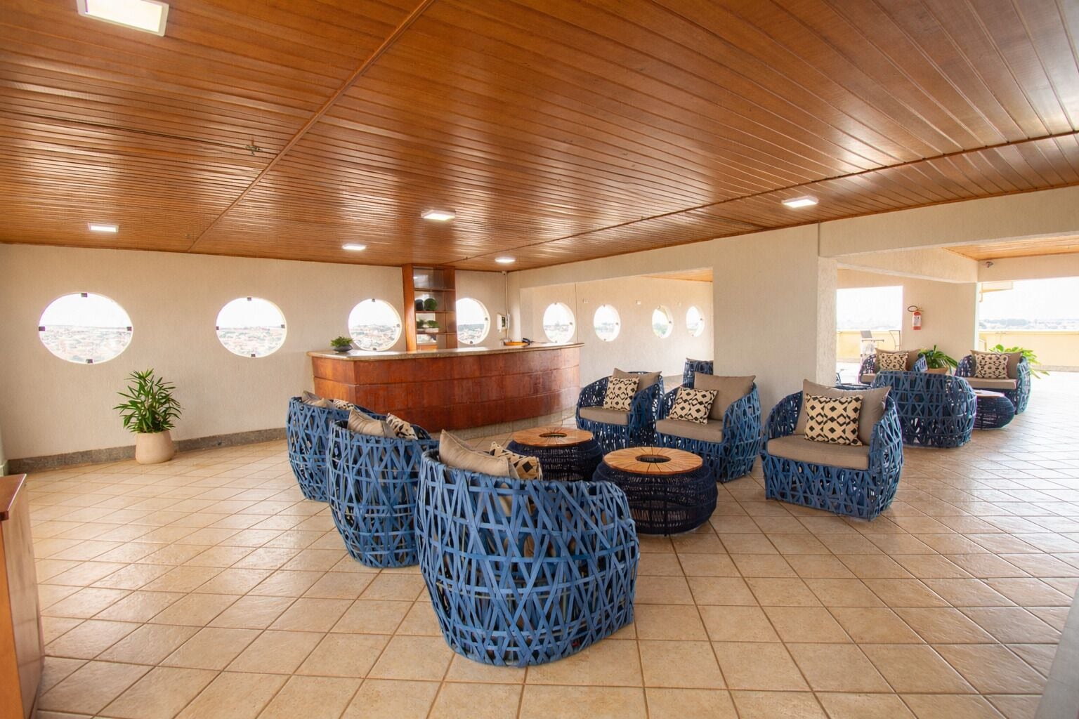 A spacious lounge area with a wooden ceiling, round seating with cushions, a central bar, and large windows offering views.
