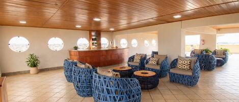 A spacious lounge area with a wooden ceiling, round seating with cushions, a central bar, and large windows offering views.