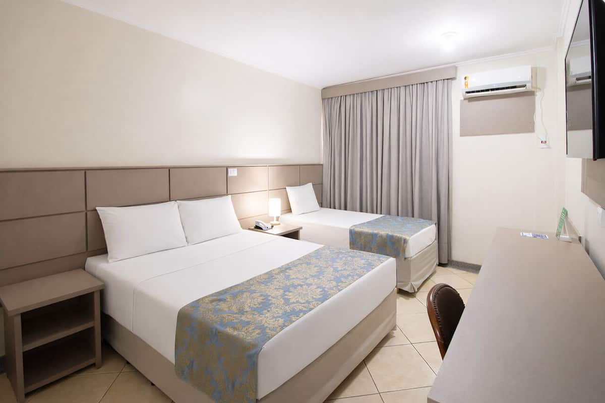 Ein Hotelzimmer mit zwei Betten, einem Schreibtisch, einem Stuhl, einem Nachttisch, einer Lampe, einer Klimaanlage und einem Spiegel.
