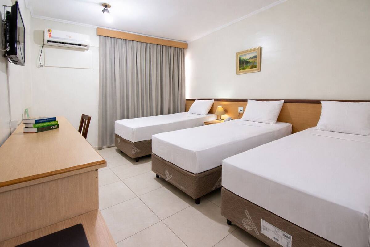 Ein Hotelzimmer mit drei Einzelbetten, einem Holztisch, einem Flachbildfernseher, einer Klimaanlage und einem Fenster mit Vorhängen.