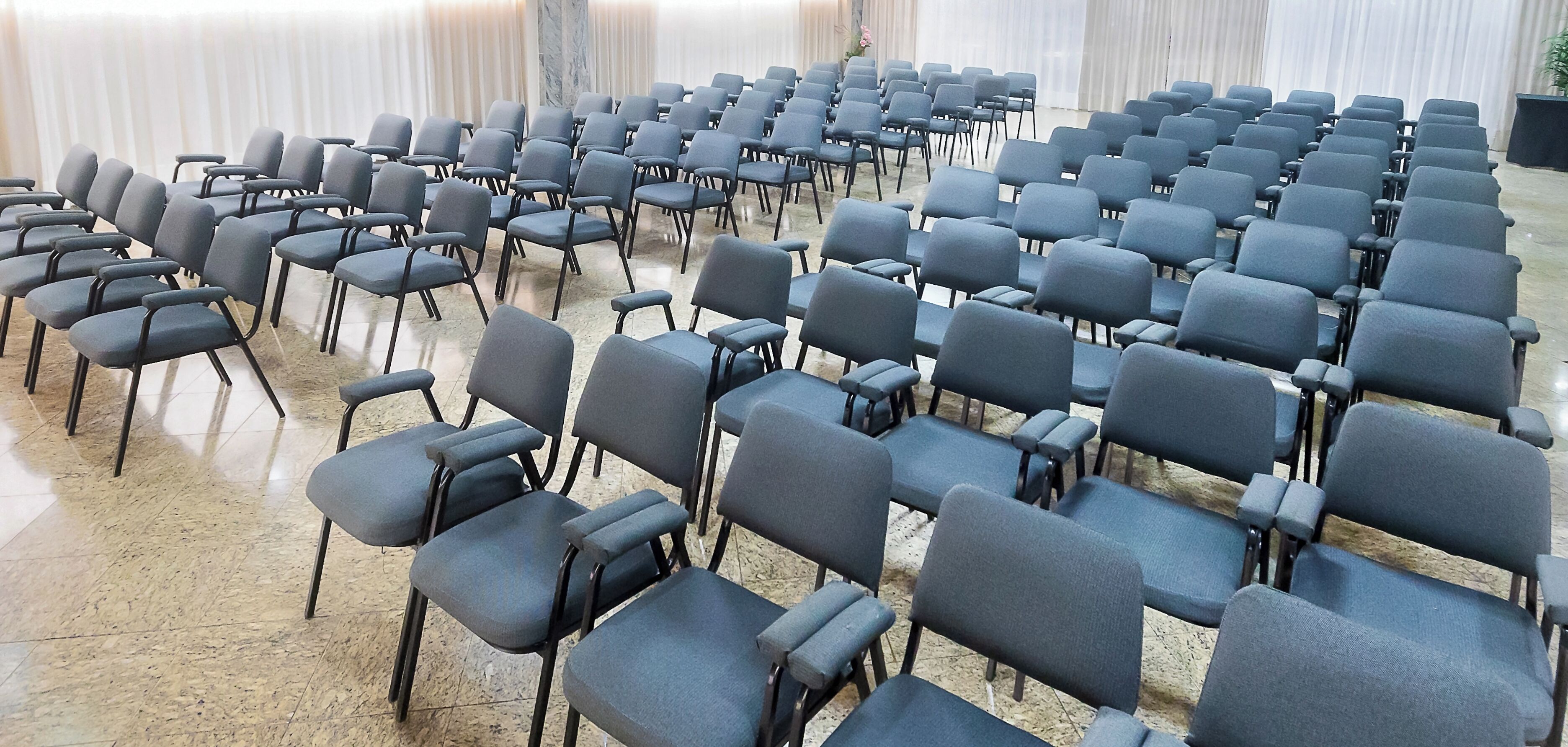 Rows of grey chairs arranged in a semi-circle in a room with a tiled floor and white curtains.