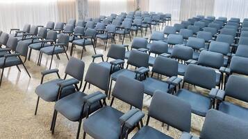 Rows of grey chairs arranged in a semi-circle in a room with a tiled floor and white curtains.