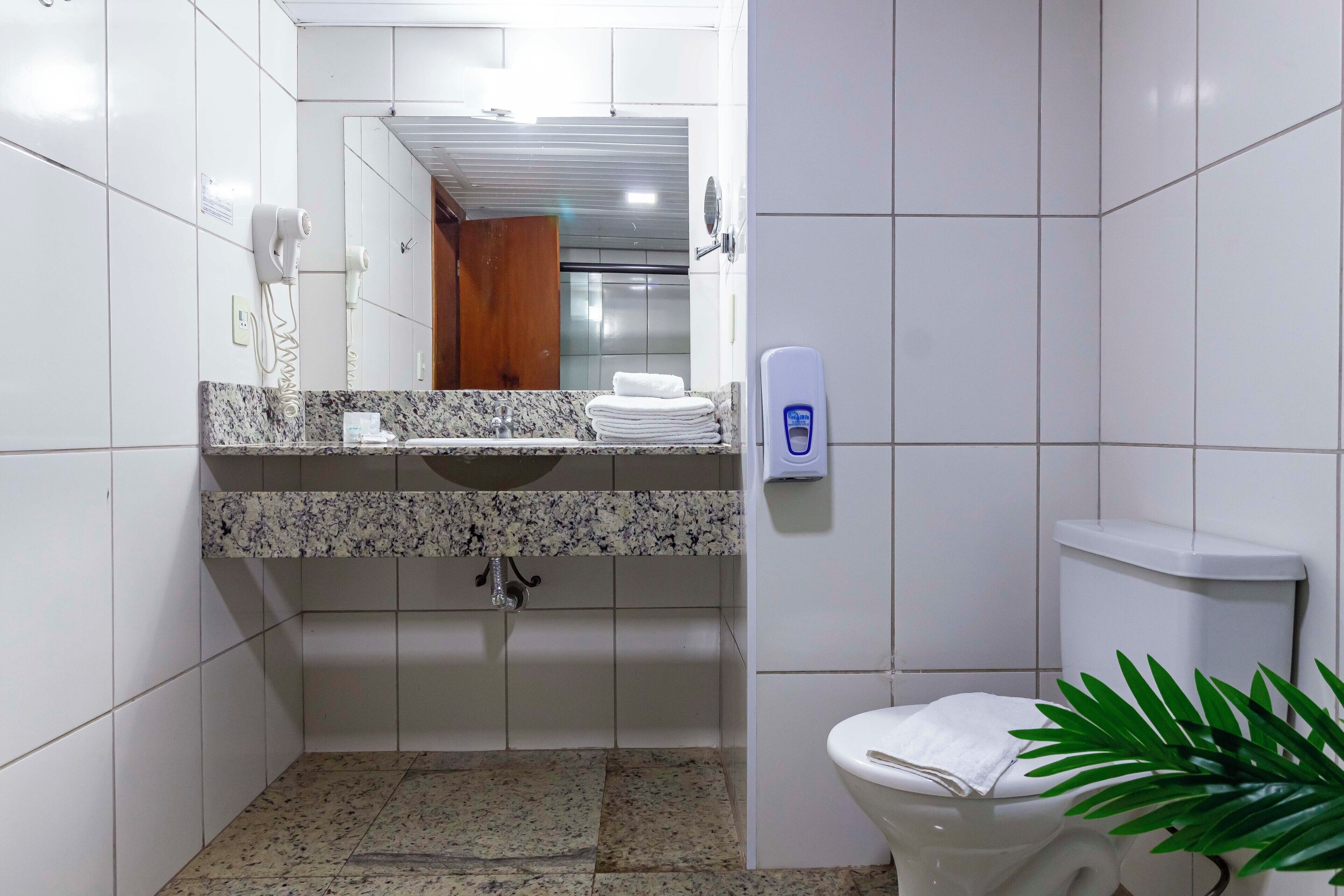 A bathroom with a marble sink, white tiles, a toilet, a hand dryer, and a plant.