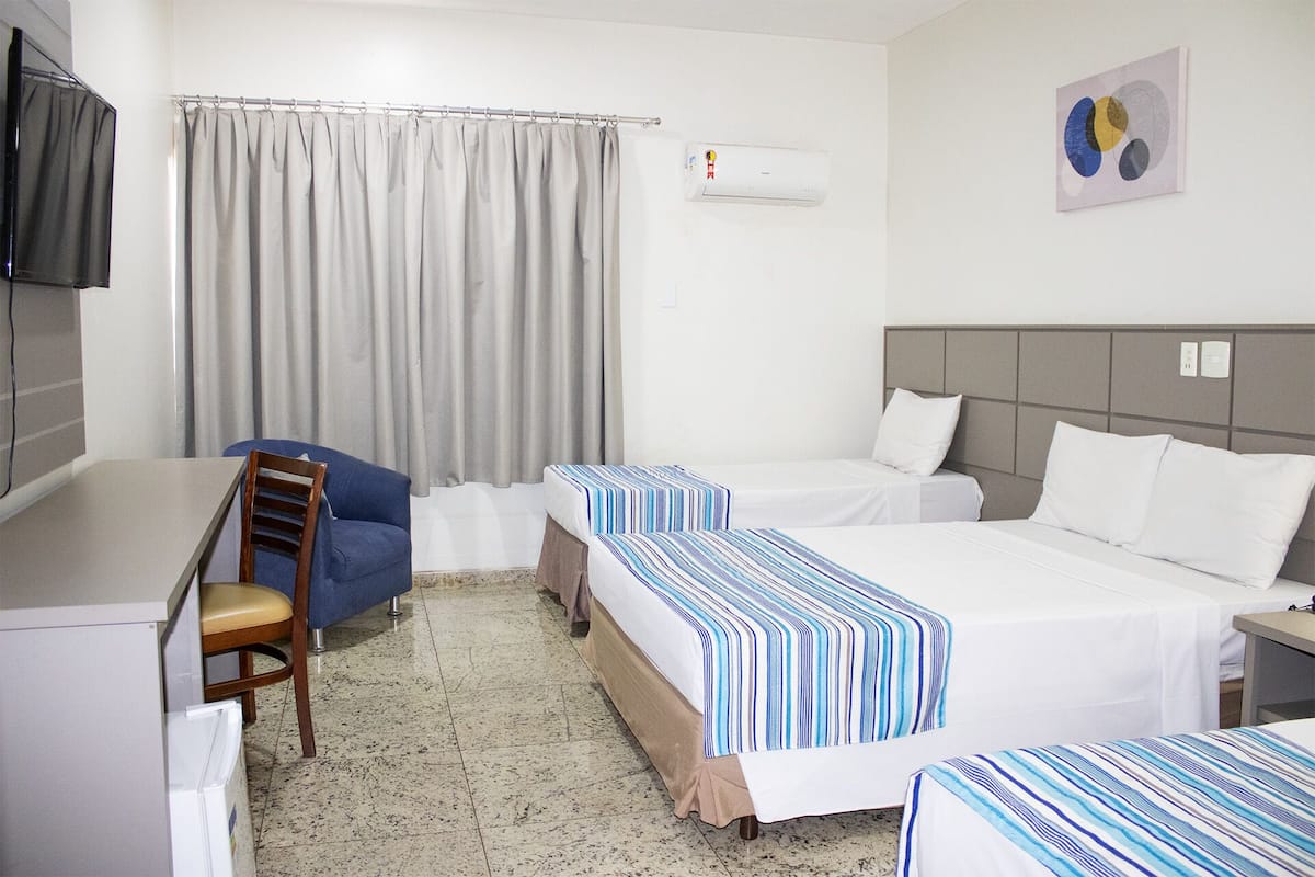A hotel room with two beds, a desk, a chair, a TV, and an air conditioning unit.
