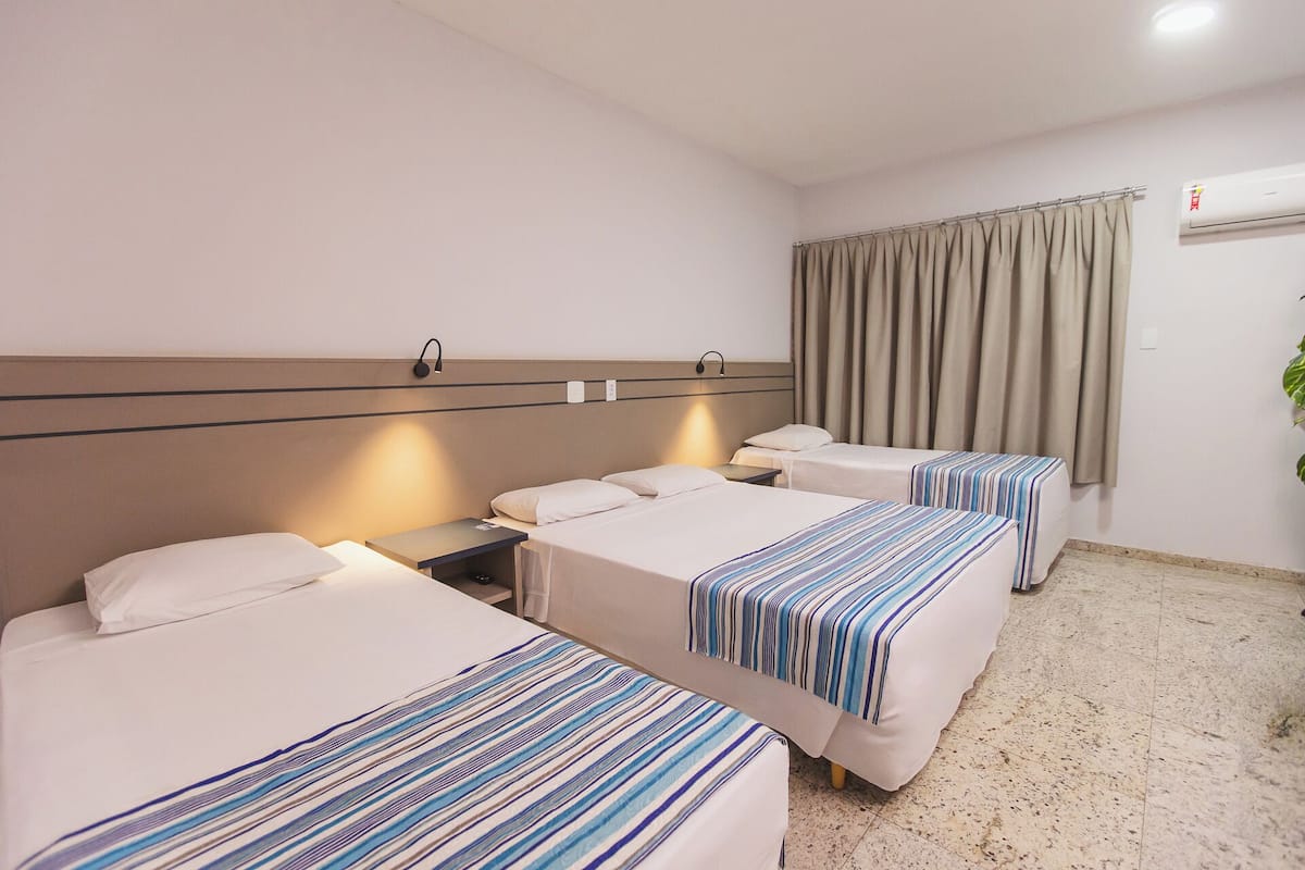 A hotel room with three beds, each with a striped bedspread, a headboard with a light fixture, and a window with curtains.