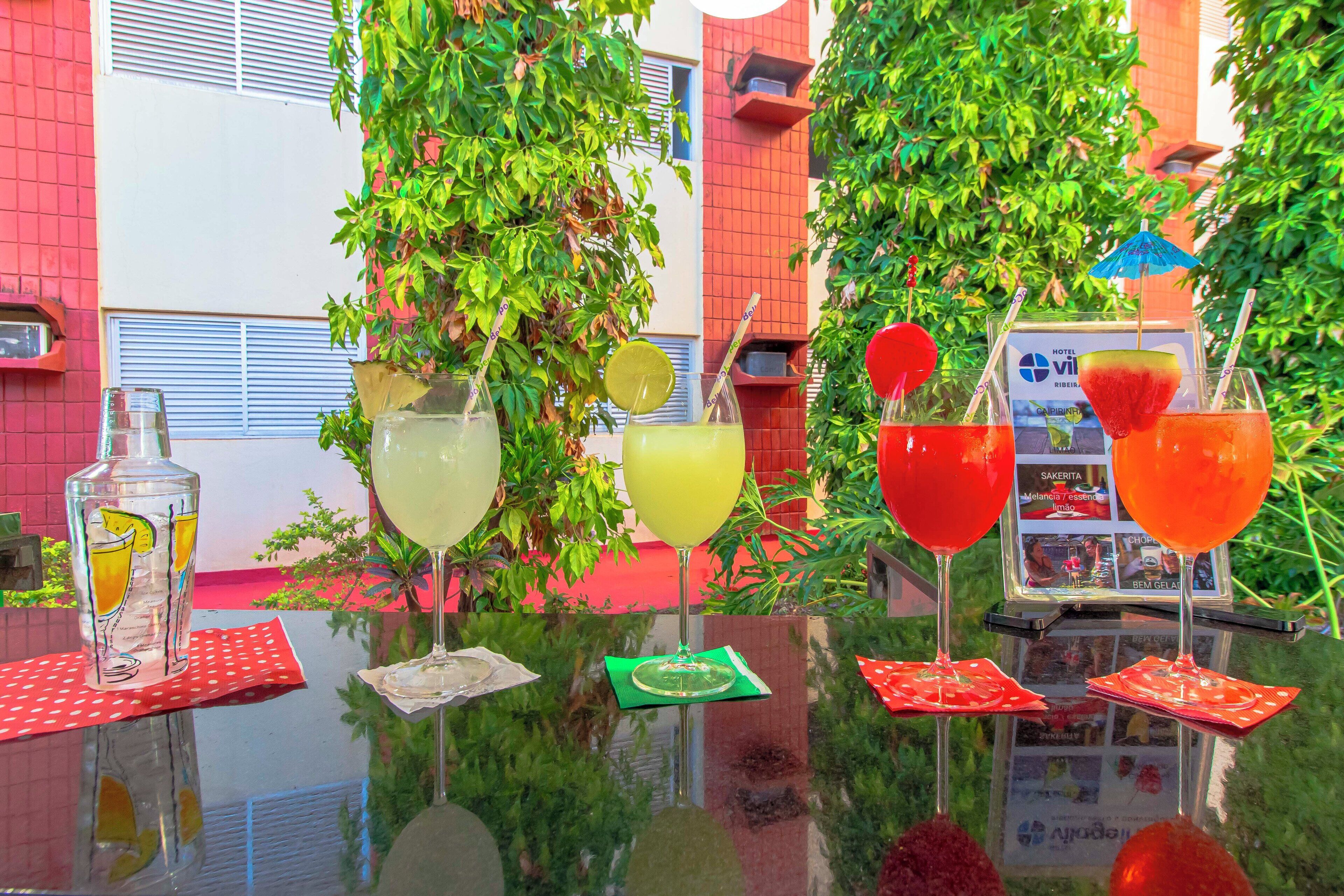 Una mesa con diversas bebidas y una botella de agua, rodeada de follaje y un muro de ladrillo rojo.
