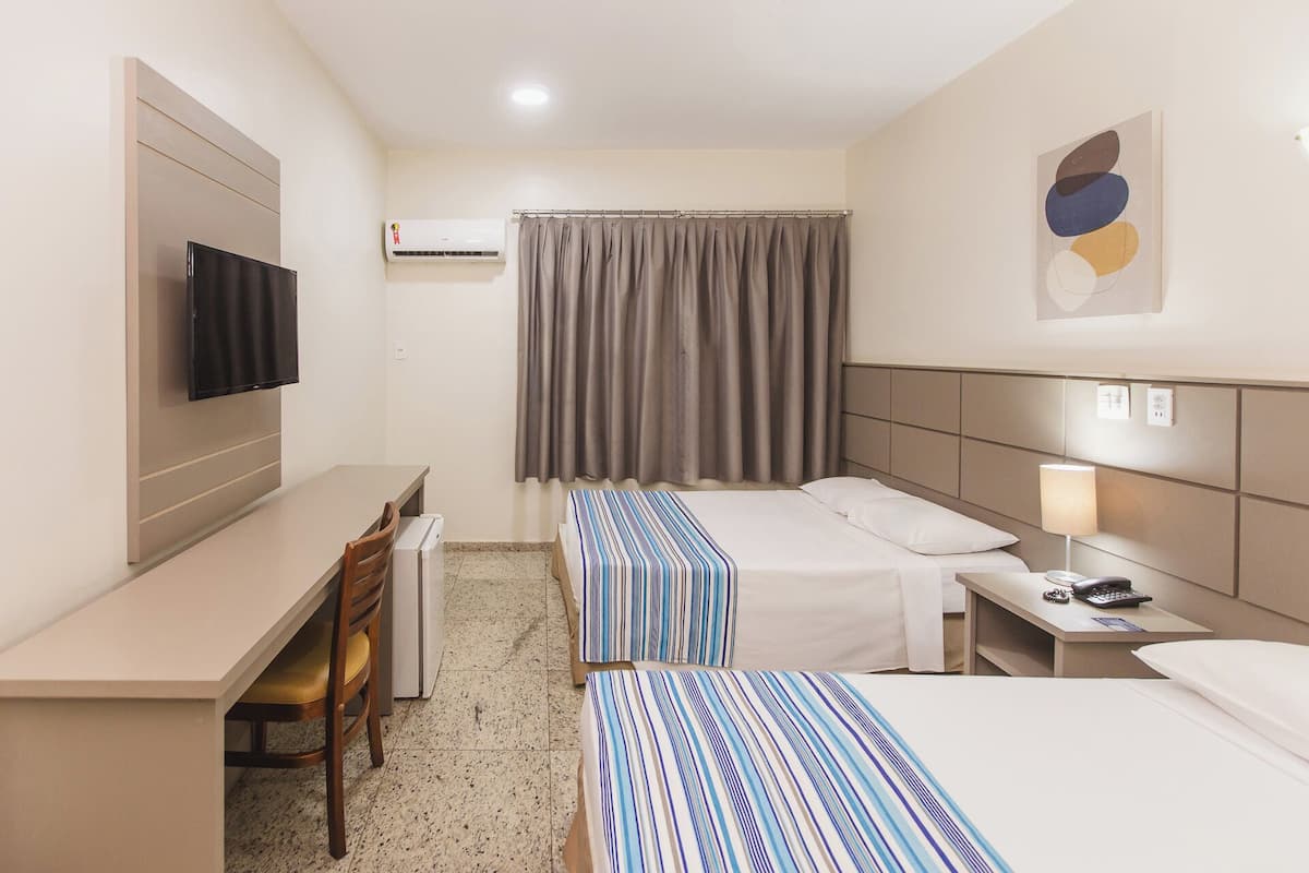 A hotel room with a bed, a desk, a chair, a television, a bedside table with a lamp, and a window with curtains.