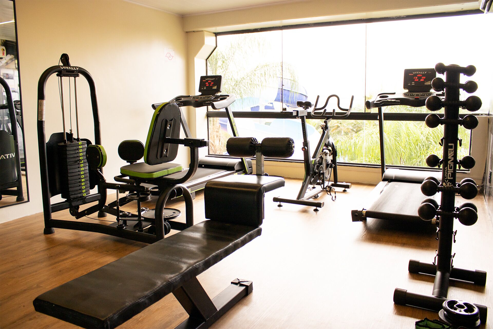 Uma academia com equipamentos de exercício, um banco e uma janela com vista para o verde.