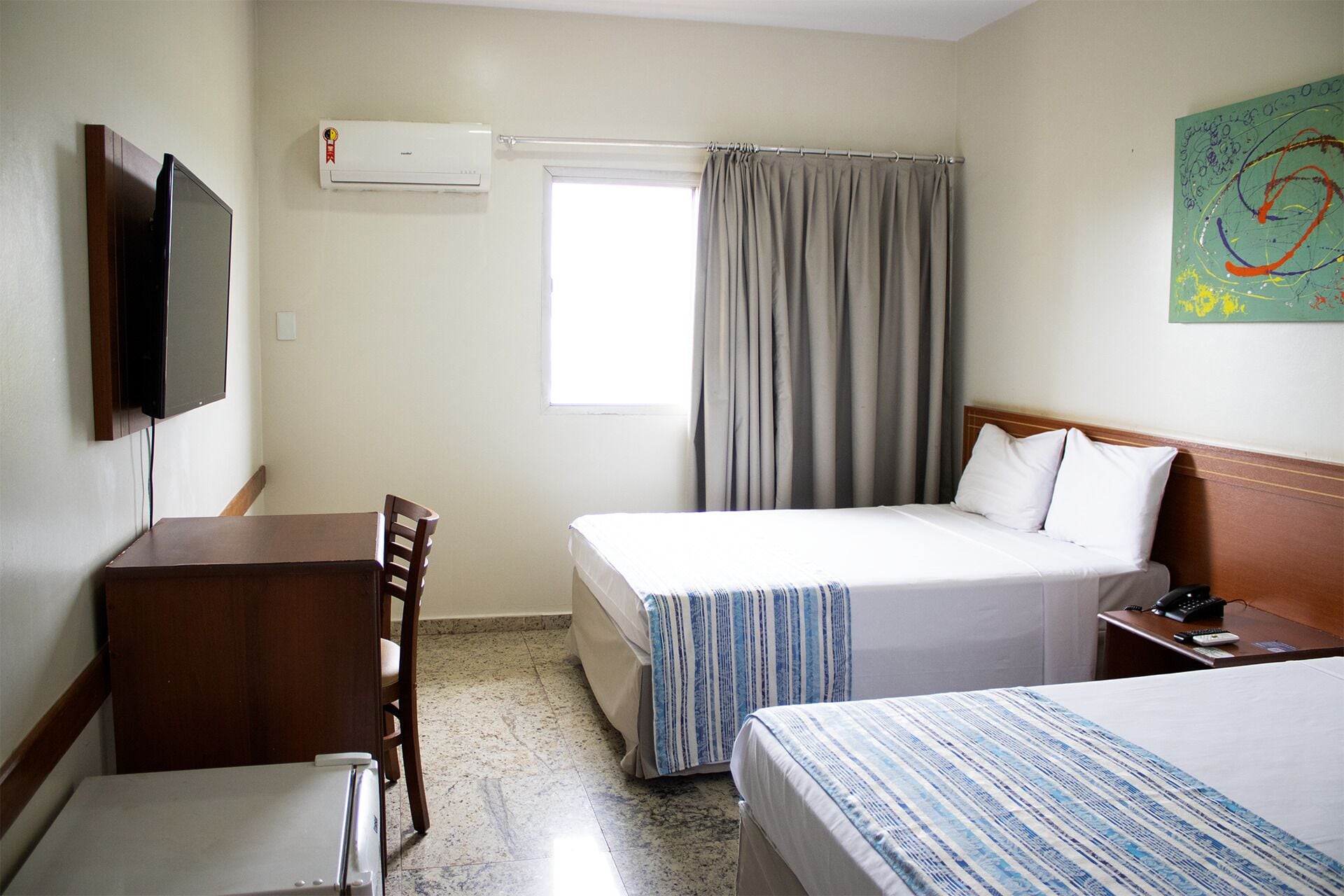 A hotel room with two beds, a desk, a chair, a television, an air conditioner, and a window with curtains.