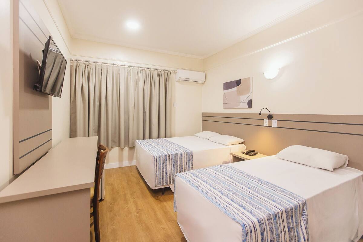 A hotel room with two beds, a desk, a chair, a TV, and an air conditioning unit.
