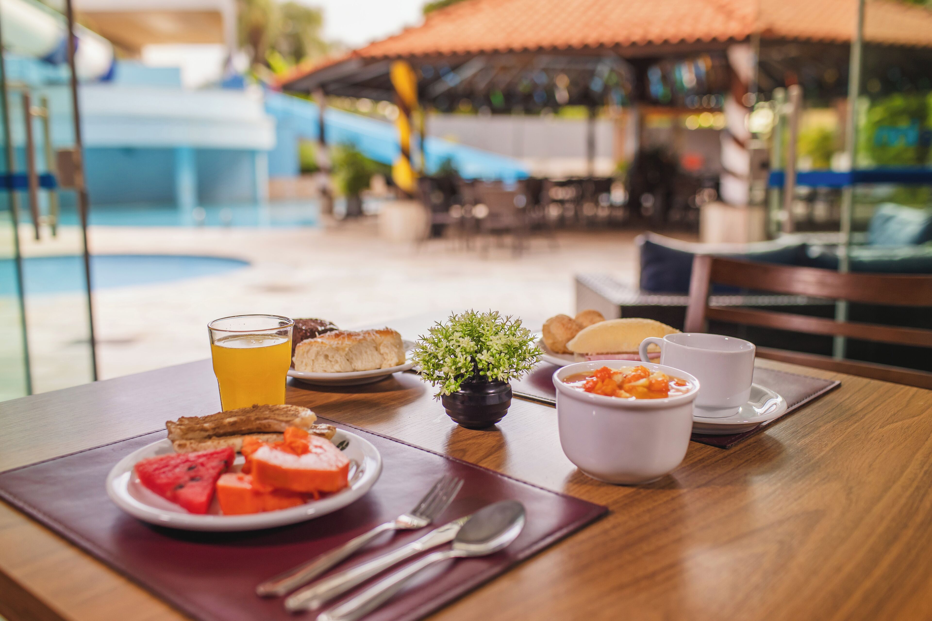 Ein Esstisch mit Frühstücksangeboten, ein Glas Orangensaft und Blick auf einen Pool sowie eine Außenlounge.