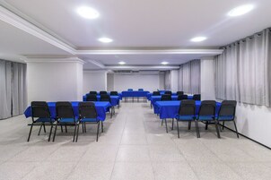 Uma sala de conferências com mesas e cadeiras dispostas em fileiras, paredes brancas e piso de azulejos.