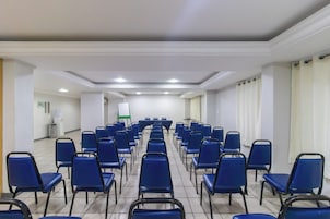 Un salón de conferencias con filas de sillas azules, una pantalla de proyector y una pizarra.