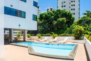 Piscina com espreguiçadeiras, cercada por vegetação e edifícios modernos.