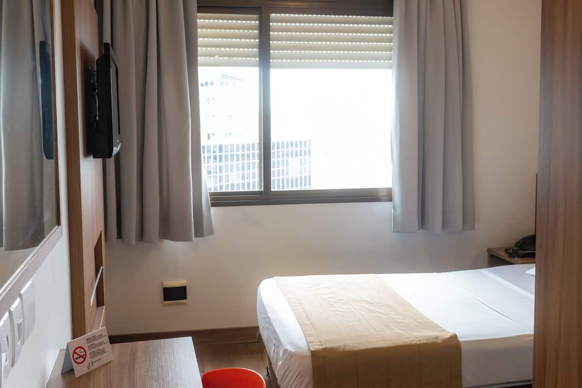 Une chambre d’hôtel équipée d’un lit, d’un bureau, d’une chaise, d’une télévision, d’une fenêtre avec des rideaux et d’un appareil de climatisation.