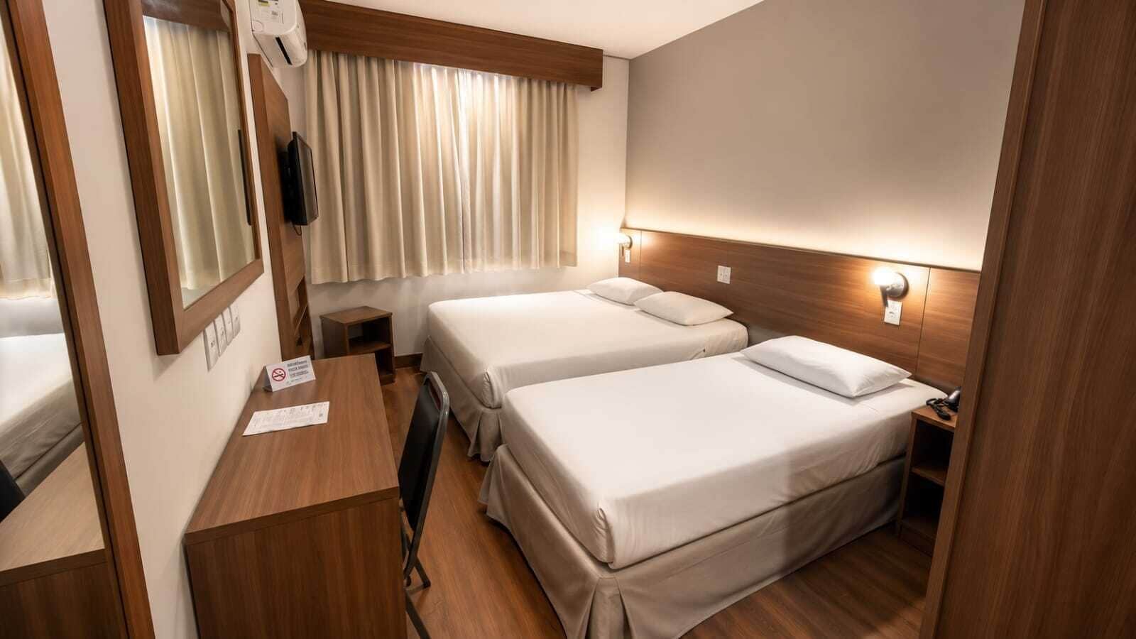 Ett kompakt hotellrum med två sängar, ett skrivbord, en spegel och ett fönster med gardiner.