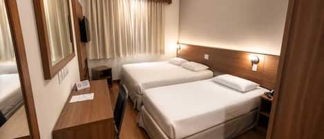 A compact hotel room with two beds, a desk, a mirror, and a window with curtains.