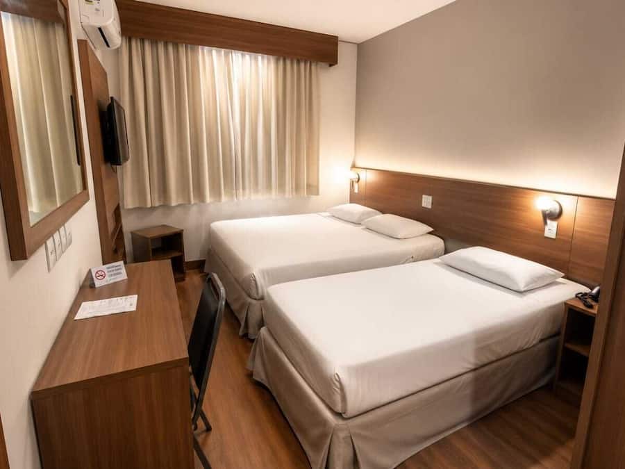 A compact hotel room with two beds, a desk, a mirror, and a window with curtains.