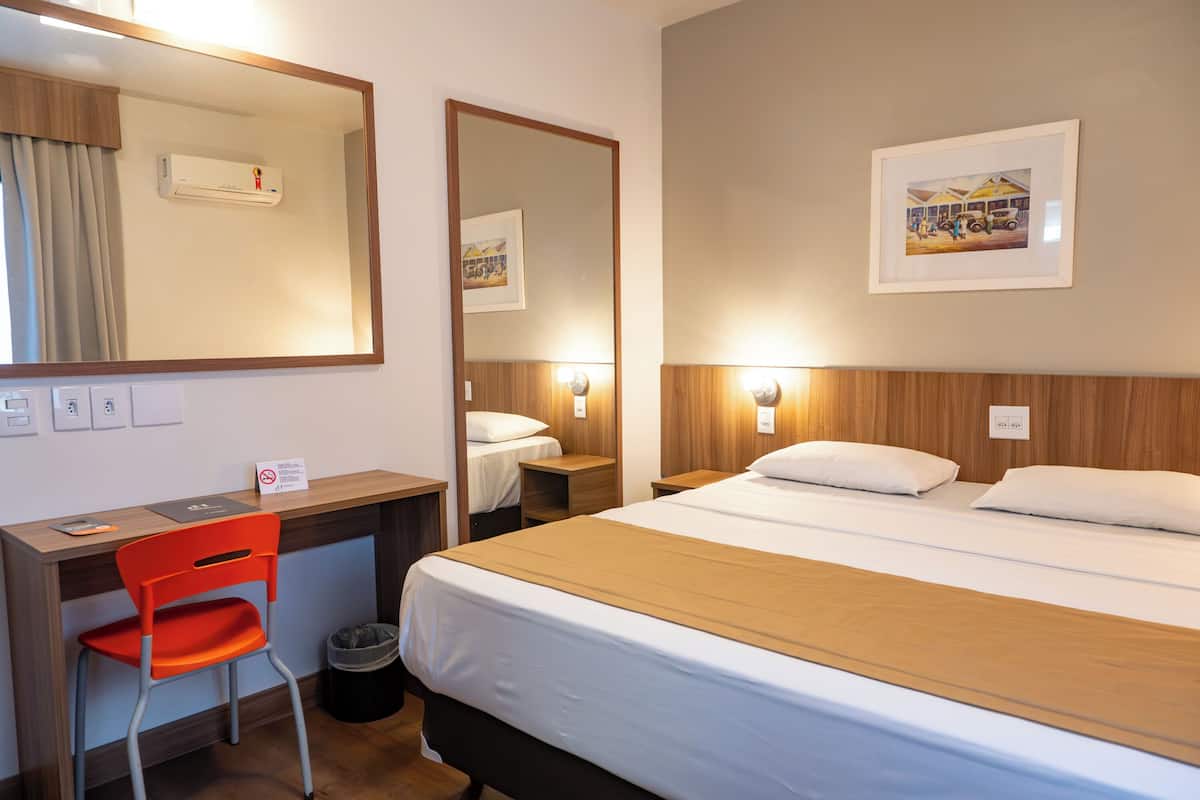 Une chambre d’hôtel comprenant un lit, un bureau avec une chaise, une poubelle, un miroir et un tableau encadré au mur.