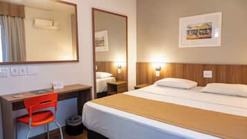 A hotel room with a bed, a desk with a chair, a trash bin, a mirror, and a framed picture on the wall.