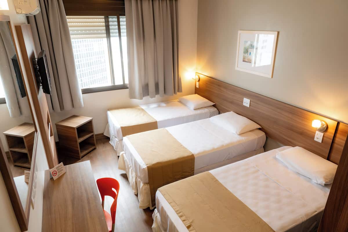 Une chambre d’hôtel avec trois lits, un bureau en bois, une chaise, une fenêtre avec des rideaux et un lampadaire fixé au mur.