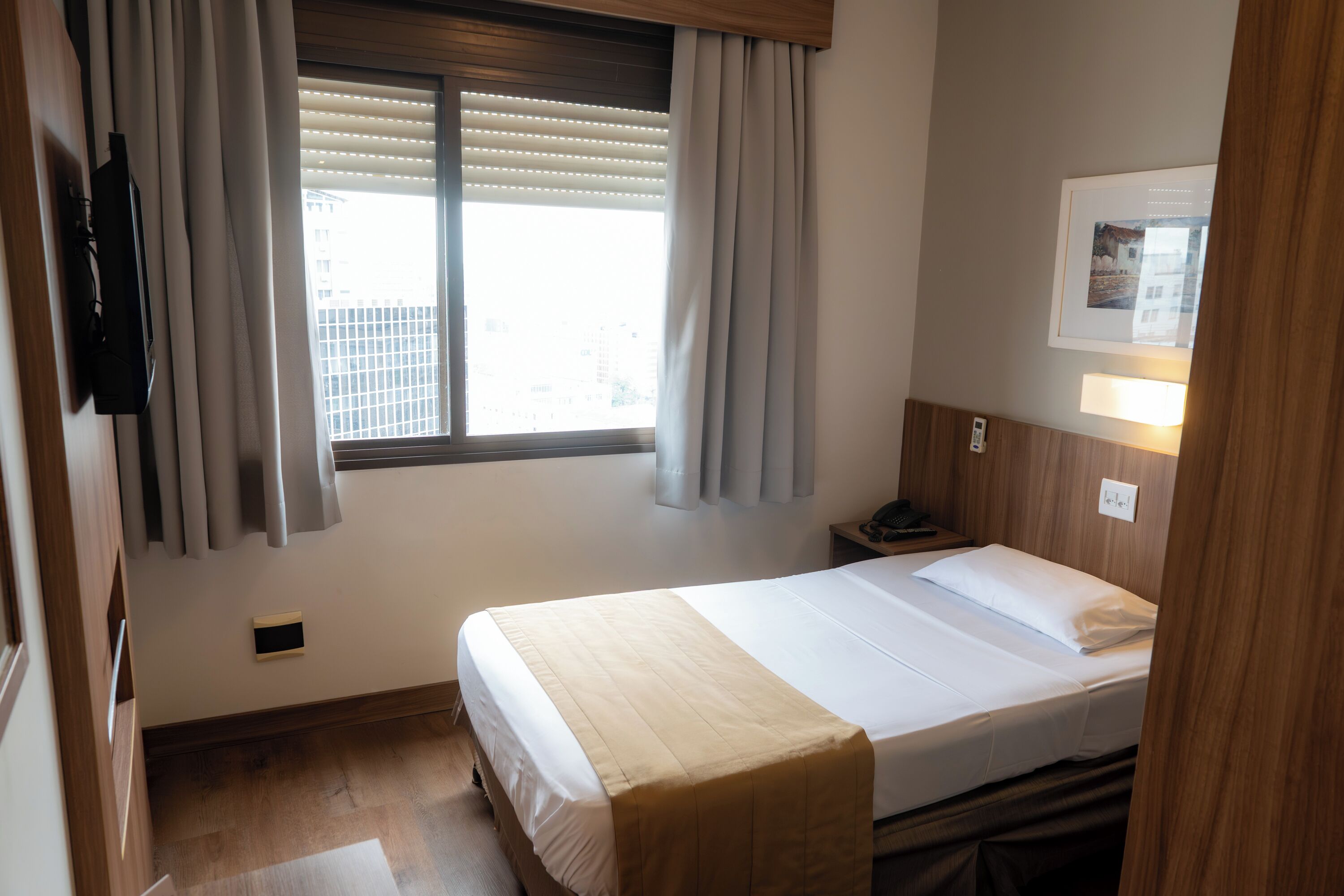 Quarto de hotel com cama, janela com persianas, mesa de cabeceira e televisão presa à parede.