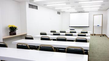 Uma sala de conferência com mesas e cadeiras brancas, uma tela de projeção e uma lousa.